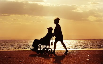Photo of sunset on beach with silhouette of a man in hat in a geriatric wheelchair being pushed by a woman figure courtesy of Pixabay