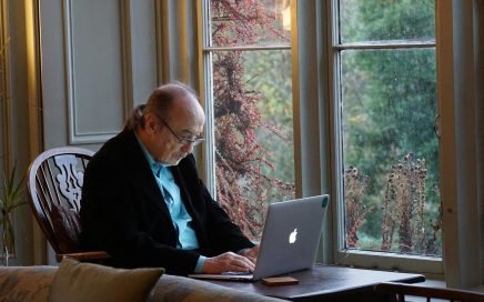 Photo of an older man sitting at a table by a window typing on a laptop courtesy Beth Macdonald on Unsplash